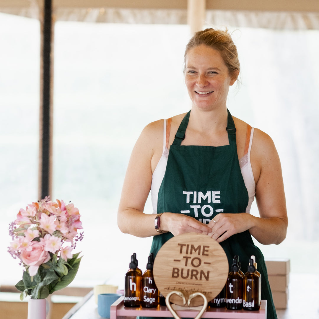 Woman in a green apron standing behind a table with bottles and a wooden sign, smiling.