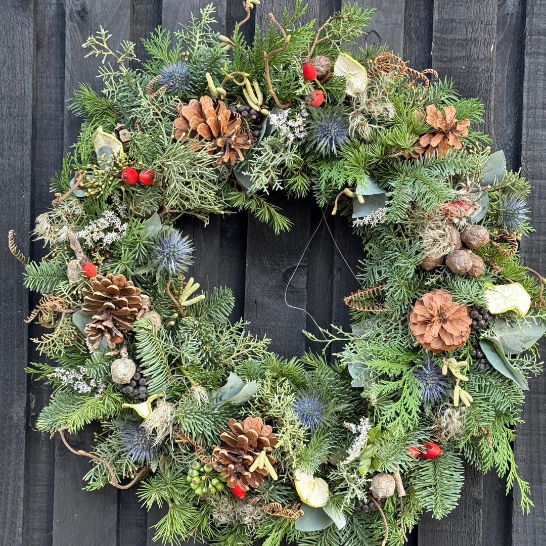 A festive Christmas wreath made of green pine and fir branches, decorated with natural brown pinecones, small red berries, blue thistle sprigs, and eucalyptus leaves, hanging against a dark weathered wooden fence.