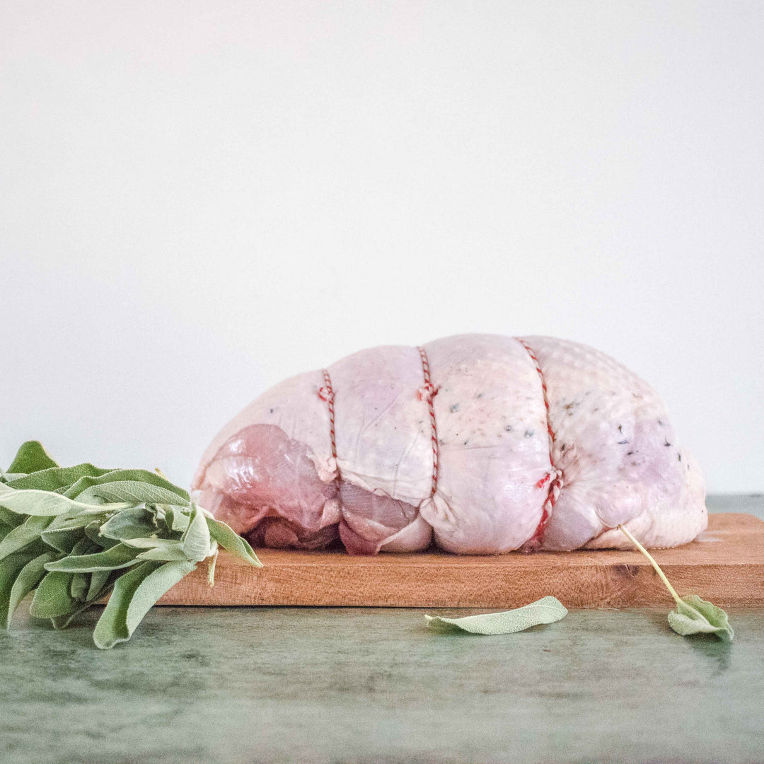 A whole, raw turkey breast meat on a wooden cutting board with a sprig of rosemary to the side, against a white background.